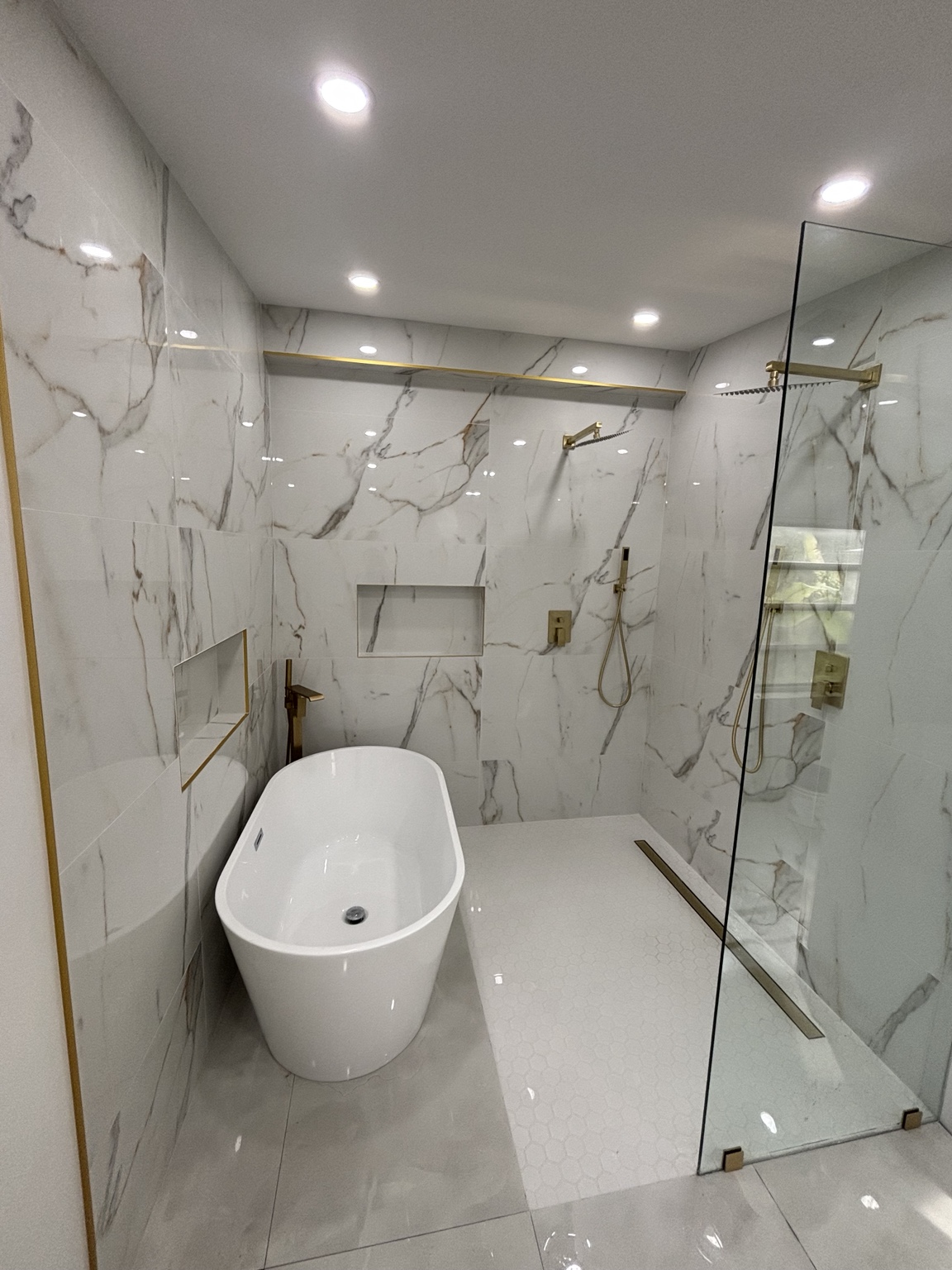 Baño terminado con tina independiente, ducha de cristal, revestimiento de apariencia mármol y accesorios dorados